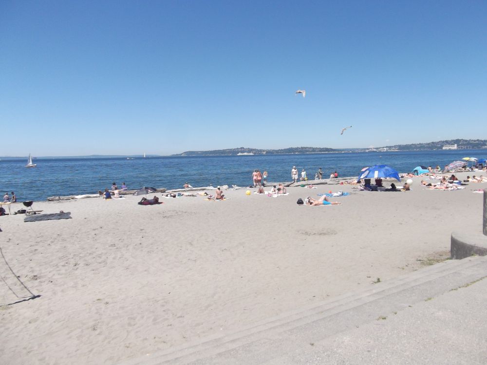Alki Beach in West Seattle on a sunny day — sandy shoreline stretching along Elliott Bay with the downtown Seattle skyline and Mount Rainier visible across the water